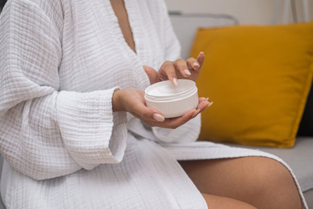 Home pampering: A woman hydrates her skin with moisturizing lotion from a jar, wearing a bathrobe, perfecting her body care.の写真素材