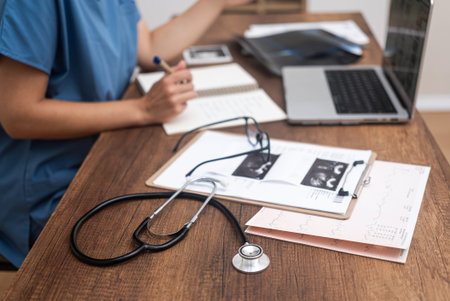 Female doctor takes notes working with patient medical documents young medic analyzes results of examinations sitting at desk with stethoscope and laptopの写真素材