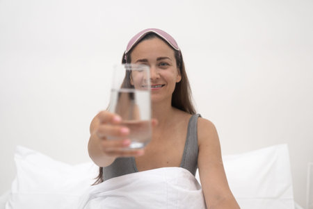 young woman, smiling, offers a glass of pure water in bed, refreshing herself after a peaceful sleep