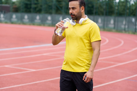 The athlete pauses on the running track, holding a transparent water bottle to rehydrate and...