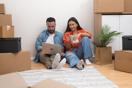 Couple sits on bare floor among cardboard boxes looking at laptop checking availability of...