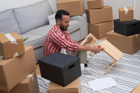 Smilling man sits with wooden furniture among various carton boxes with stuff for interior decoration enjoying moving in new contemporary house in another townの写真素材