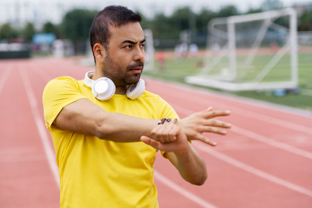 Thoughtful sportsman with earphones warming up arms muscles preparing body for training on running track at city sports ground with soccer arena in summerの写真素材