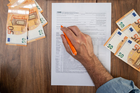 Agricultural worker completes 1040 tax form with pen in hand detailing seasonal earnings and travel expenses to determine tax liability sitting at table with euro billsの写真素材