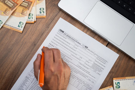 New homeowner carefully completes 1040 form with pen reporting income and mortgage interest to calculate tax obligations sitting at table with euro bills and laptopの写真素材