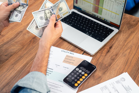 Businessman counts dollar banknotes over utility bills and laptop working with digital spreadsheets...
