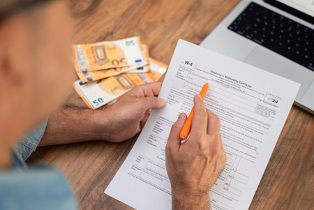 Man filling out W-4 tax form with euro banknotes and laptop on wooden desk. Financial management and tax preparation processの写真素材