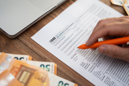 Close-up of a hand filling out a 1040 tax form with euros and a laptop on a wooden desk. Tax filing and financial preparation concept, ideal for accounting and international finance themesの写真素材