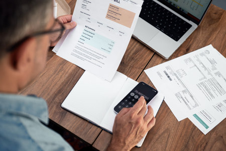 Man calculating utility expenses using a smartphone at a wooden desk. Household finance management and budget planning concept.の写真素材