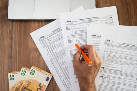 Close-up of a person filling out tax forms on a wooden desk with an orange pen. Euro bills and a laptop are nearby, representing financial calculations and tax preparation. tax season, accountingの写真素材