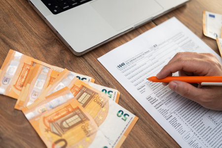 Hand filling out a 1040 tax form with euros and a laptop on a wooden desk. Tax preparation and international finance concept, ideal for accounting and tax season themesの写真素材