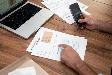 Close-up of a person using a smartphone calculator while reviewing utility bills on a wooden desk. Household expenses and financial management concept. Budget planning and bill payments.の写真素材