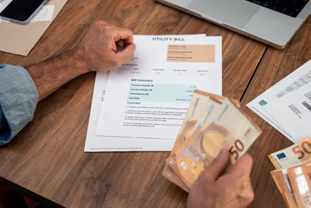 Man analyzing utility bill while holding euro banknotes at home. Focus on household expenses, tax management, and online payment solutionsの写真素材