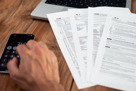 Hand using a smartphone calculator next to various tax forms on a wooden desk. Tax season, accounting, and financial planning conceptの写真素材