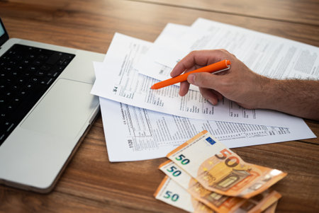 Hand filling out tax forms on a wooden desk beside a laptop and euro bills. Tax preparation and financial documentation in progress. accounting, financial planning, and tax season.の写真素材