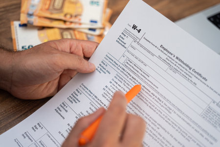 Close-up of a mans hands filling out a W-4 tax form with an orange pen, with euro banknotes in the background. Tax filing and financial management concept during tax seasonの写真素材