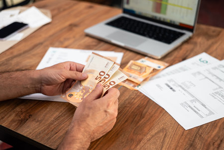 Man counting euro banknotes while managing household expenses and utility bills. Focus on financial planning, tax calculations, and online payment managementの写真素材