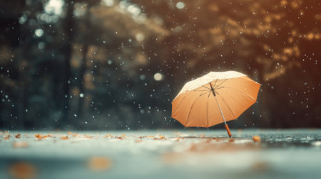 Rain falling on wet ground, creating puddles among fallen leaves, with an orange umbrella standing prominently. Trees in the background blur softly, enhancing the autumnal atmosphereの素材