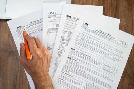 Close-up of a man filling out tax forms with an orange pen at a wooden desk. Personal finance and tax preparation concept during tax seasonの写真素材
