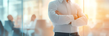 Businessman standing confidently with arms crossed in a bright, modern office while colleagues engage in a meeting, embodying leadership and success in the corporate worldの素材