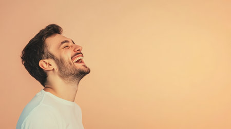 Cheerful young man with a beard laughing with closed eyes and displaying perfect white teeth against a beige background, radiating joy ideal for dental and cosmetic themesの素材