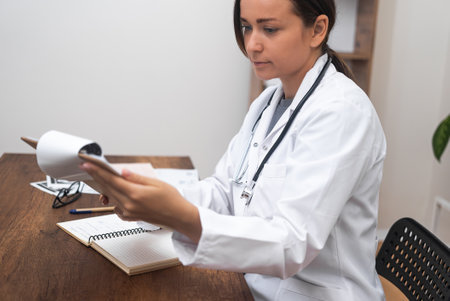 Woman doctor diagnoses patient studying images of examination and medical conclusions attentive therapist in medical coat sits at table in office in hospitalの写真素材