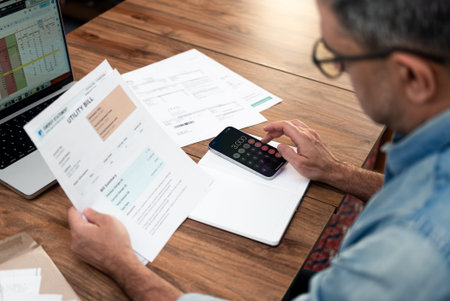 Senior man counts total sum of utility bills with mobile app at wooden table, mature male accountant checks household expenditures for current month, entrepreneur plans budgetの写真素材