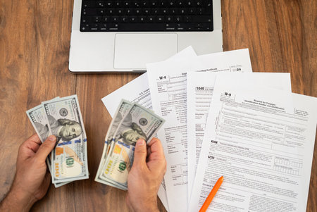 Male entrepreneur holds dollar banknotes in hands counting cash for filling different tax forms in accordance with strict requirements sitting at workplace with laptopの写真素材