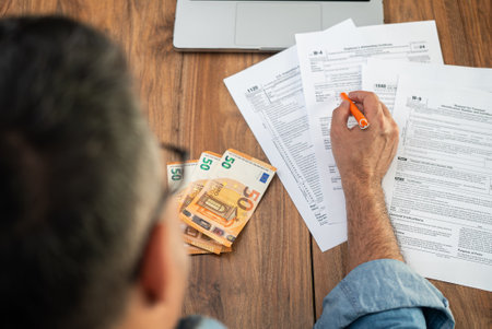 Man accountant examines different types of tax forms for businesses to avoid penalties from government at workplace with euro bills and laptop in officeの写真素材