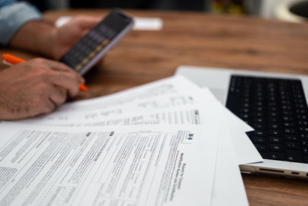 Man uses calculator on smartphone to calculate taxes preparing to fill different tax forms reporting annual income ensuring compliance with tax laws at desk with laptopの写真素材