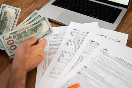 Male entrepreneur holds dollar banknotes counting cash for indicating additional income source in tax forms sitting at workplace with notebook in officeの写真素材