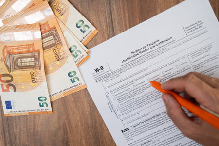 Independent contractor sits at wooden table with euro bills with pen in hand filling W-9 form to...
