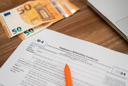 Euro bills lie scattered near essential W-4 form on table alongside pen and laptop as worker prepares to fill out form to ensure correct federal tax deductionsの写真素材