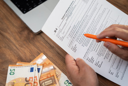 Male remote contractor completes W-4 tax form with euro bills scattered on table at home to ensure being taxed correctly for federal income by employerの写真素材