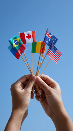 Hands holding small flags of different countries against a vibrant blue sky, representing international cooperation, diversity, and global communityの素材