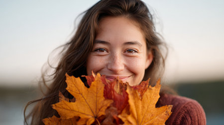 Happy young woman smiling while holding a vibrant bunch of colorful maple leaves in front of her face, enjoying the beauty of autumn in a sunny park settingの素材