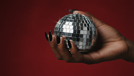 Womans hand with long black stiletto nails gently holding a sparkling disco ball against a vibrant red background, representing retro party, celebration, glamor, and fashionの素材
