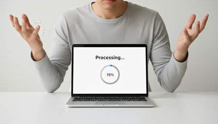 Man feeling frustrated with a slow laptop displaying a processing... Message and 15 percent loading progress on the screen, showing helplessness with raised handsの素材