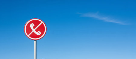 Red and white no cell phone sign, featuring a phone handset with a prohibitive line through it, indicating a restriction or forbidden use of mobile devices against a clear blue skyの素材