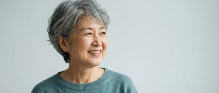 Happy senior asian woman with short grey hair smiling and looking to the side, conveying aging gracefully, confidence, warmth and optimistic wellbeing on a white studio backgroundの素材