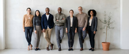 Diverse group of smiling professionals standing together against a light gray wall, representing inclusion and a collaborative team environment in a modern office spaceの素材