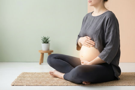 Pregnant woman seated cross legged on a mat, hands cradling her baby bump in a calm, mindful prenatal yoga pose with a plant in the background, radiating wellness and anticipationの素材