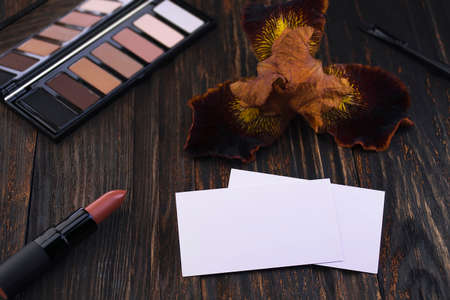 Business cards, pink matte nude lipstick and iris flower on a wooden table. A brown eyeshadow palette and a brush in the background. Cosmetics, trendy glamorous colorの写真素材