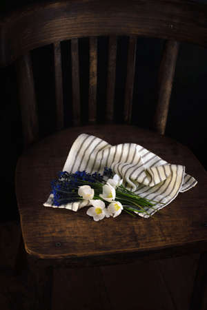 Bouquet of spring white and blue flowers wrapped in light striped fabric on a vintage wooden chair. Dark background. Top view. Romantic gift for print, cozy moodの写真素材
