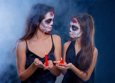 Mom and daughter with a sugar skull make-up on a dark background are holding candles in their hands....