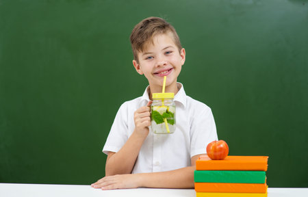 The boy has lunch at school. Against the background of a green school board, a stack of books is standing nearby. Boy drinking lemonadeの写真素材