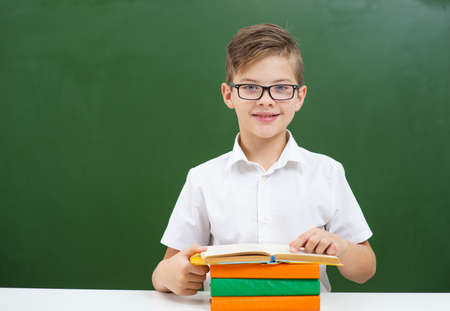The boy in glasses is engaged in school, sitting on the background of a green blackboard and reading a bookの写真素材