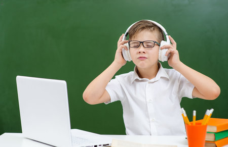 A boy with glasses is engaged in school, sitting on the background of a green board at a laptop and listening to music on headphonesの写真素材