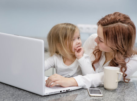 Teen girl works at a laptop, looks at her little sister and smiles at her, sitting at home in the...