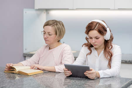 A teenage girl sitting with a tablet computer in her hands and in headphones, next to her mother is sitting in glasses and reading a book. In the kitchen in your homeの写真素材